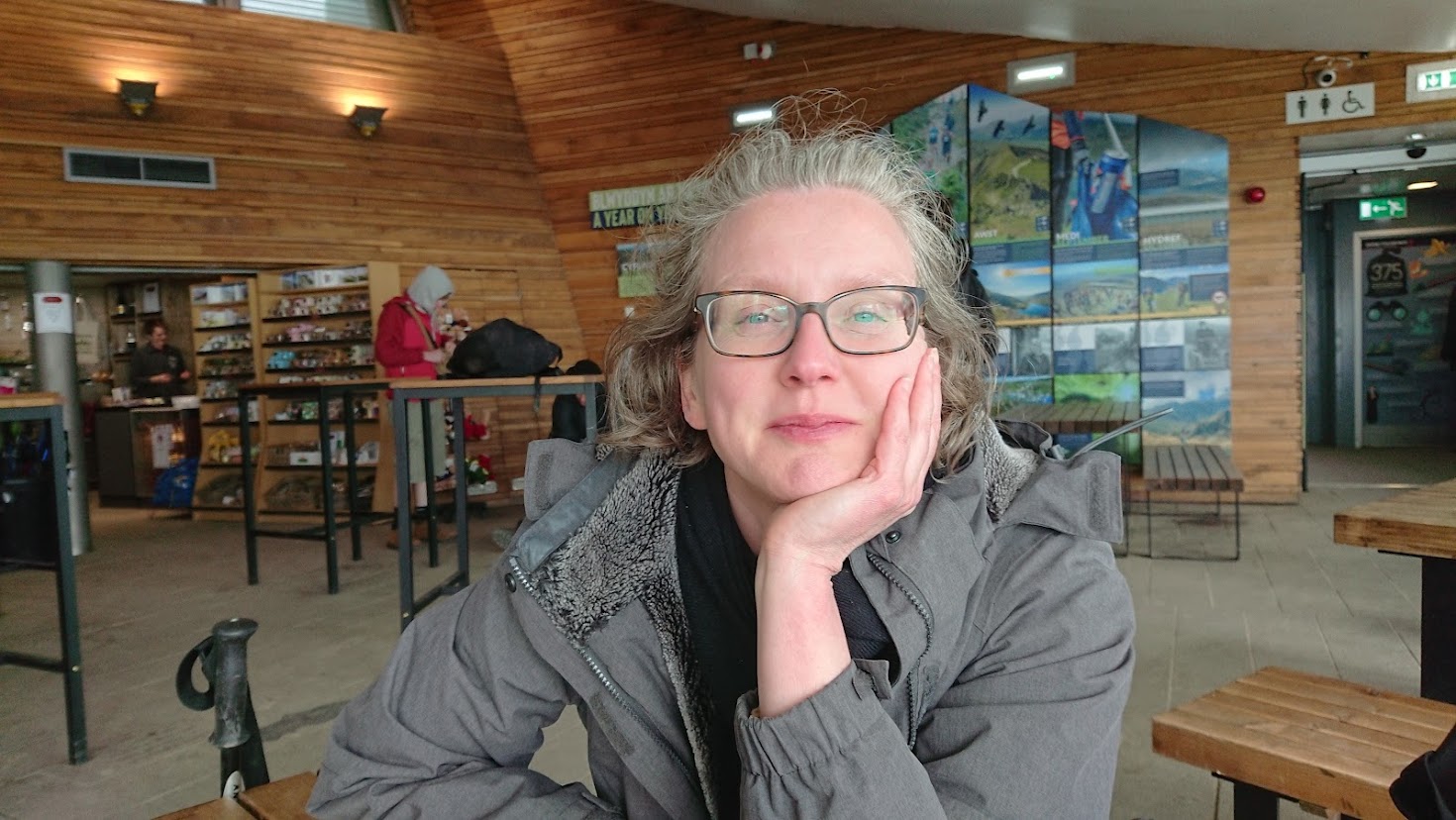 Sally is sitting at a table with one hand cupping her chin, smiling towards the camera, in the cafe at the top of Snowdon, Wales, with the small outdoors equipment shop in the background behind her