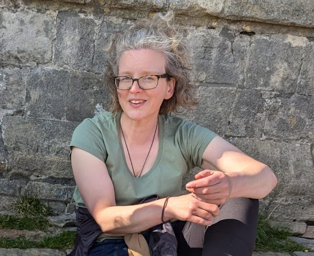 Sally sitting smiling against a grey stone wall (part of Glastonbury Tor), hair blowing in the wind